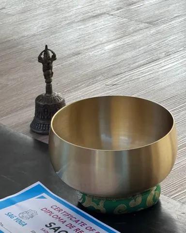 Un cuenco tibetano sobre su almohadilla de seda verde, junto a una campana ritual y un diploma de SAG Yoga, sobre una mesa de madera clara.