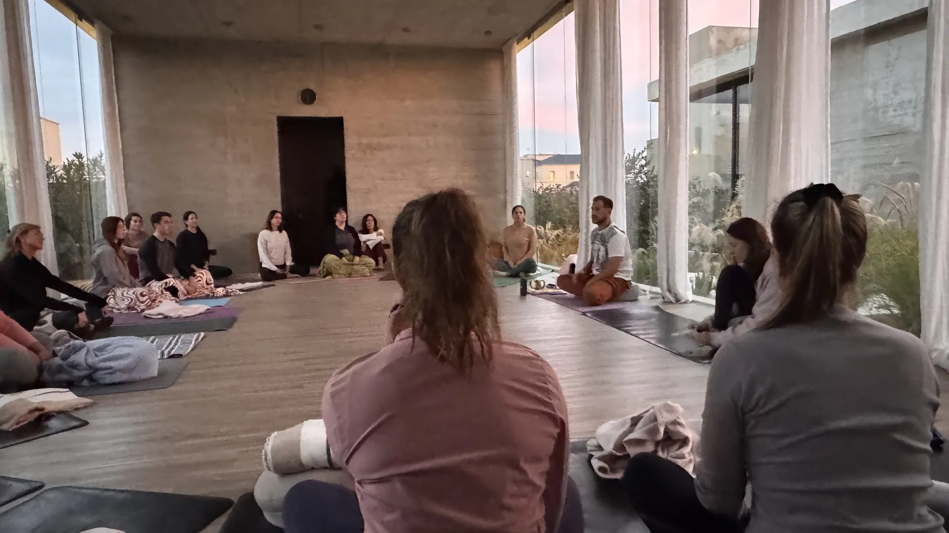 Un grupo sentado en ronda, meditando al atardecer en una sala de hormigón con ventanales del piso al techo.