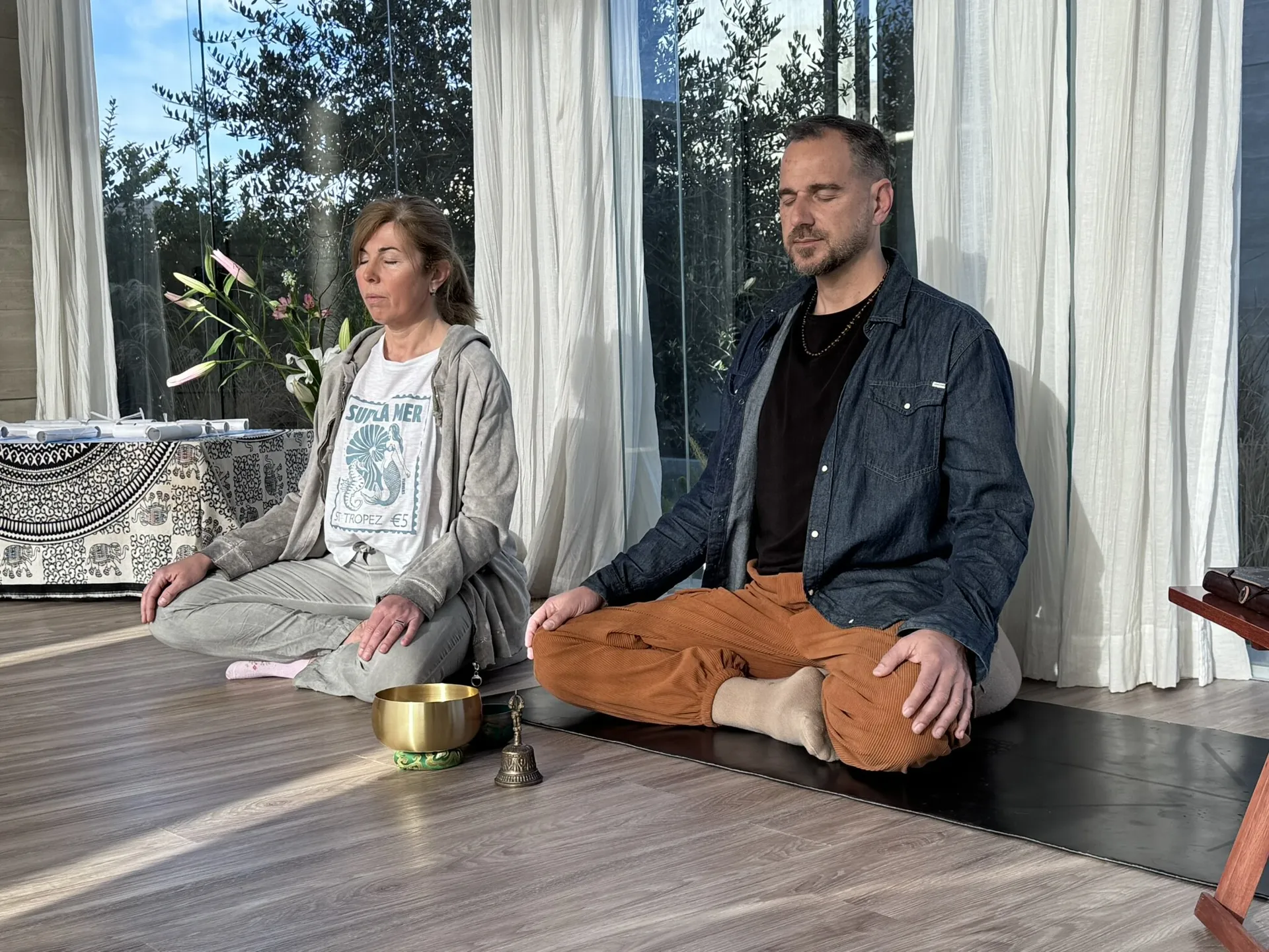 Marcelo Poppi y una practicante sentados en el piso en postura de meditación, con los ojos cerrados, frente a un ventanal con cortinas claras; delante de ellos un cuenco tibetano y una campana.