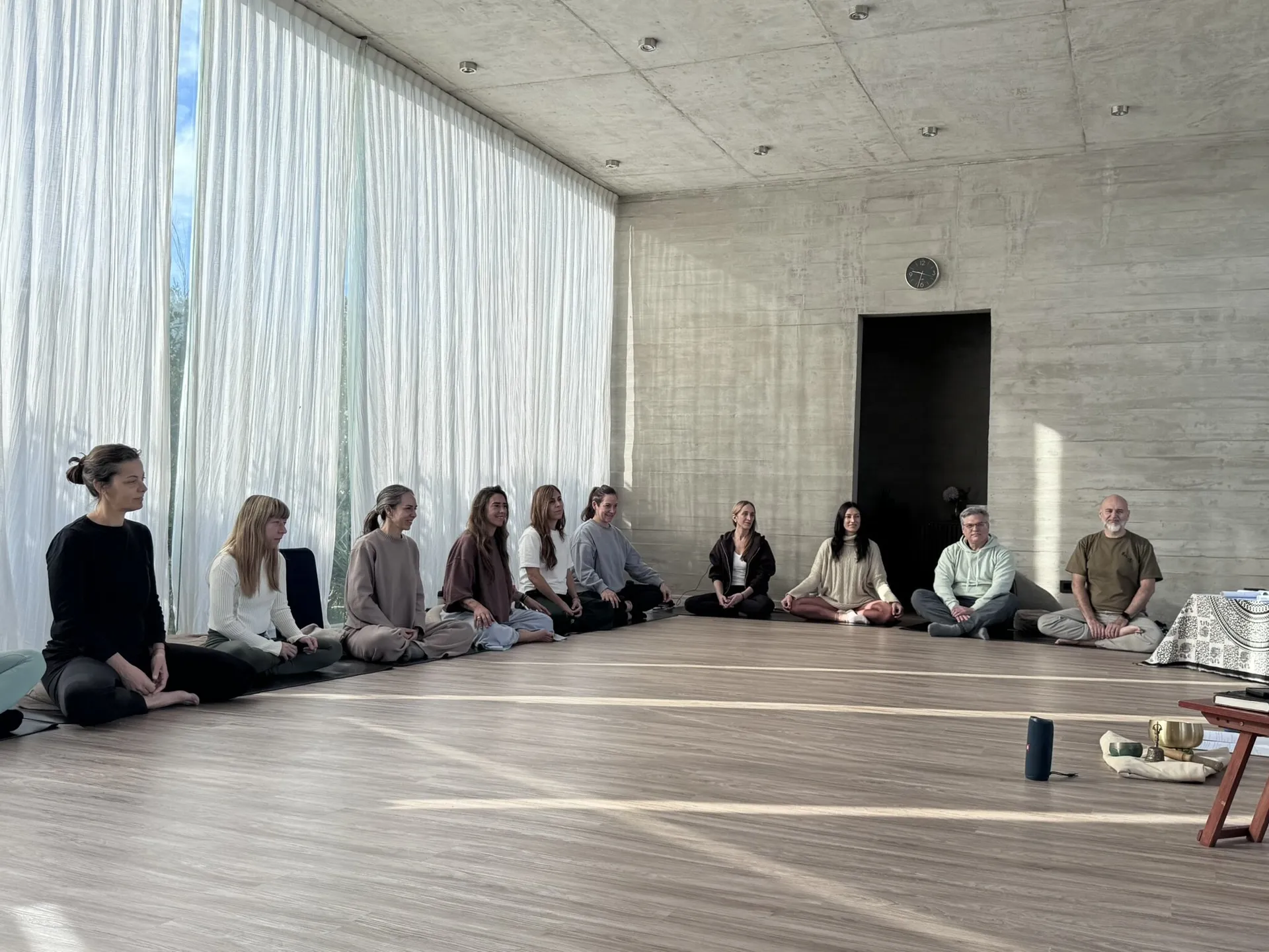 Un grupo sentado en ronda sobre el piso de madera de una sala de hormigón, en postura de meditación, con un ventanal de cortinas claras a un costado.